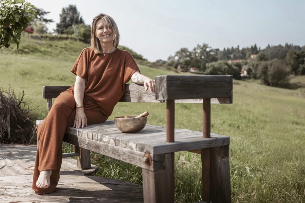 Portrait de Laurence Rey assise sur un banc en bois, avec son bol tibétain posé à proximité, entourée de verdure.
